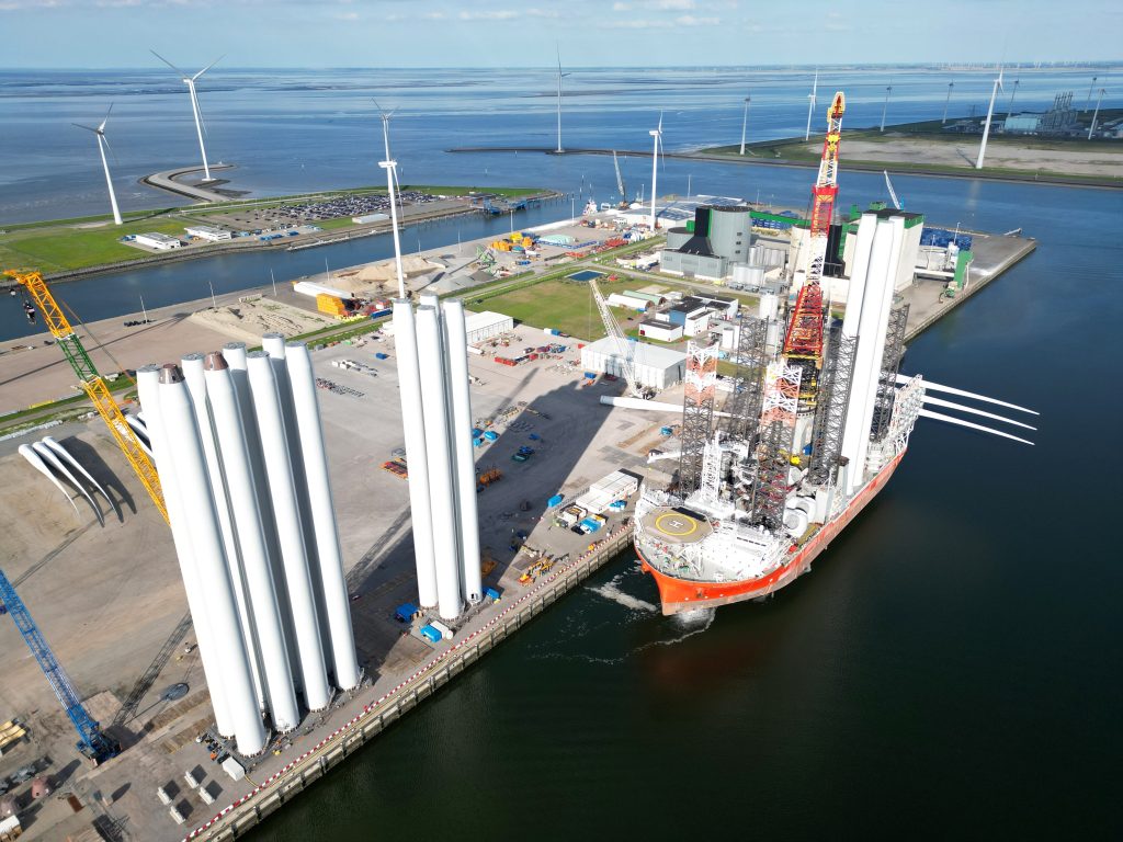 Opslag en transport van windturbine-onderdelen in een industriële haven met operationele windmolens op de achtergrond