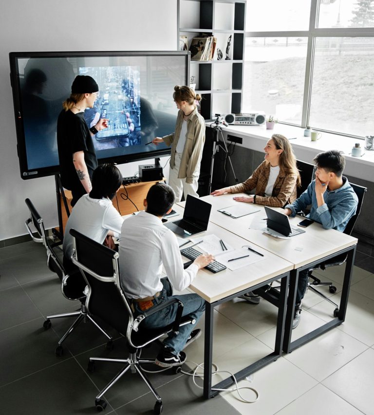 Team sits around a table in discussion with screen presentation in the background.