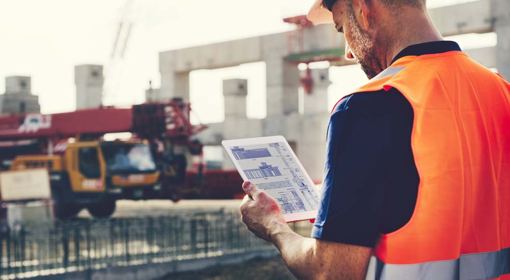 Man op bouwplaats met oranje hesje kijkt naar bouwtekening