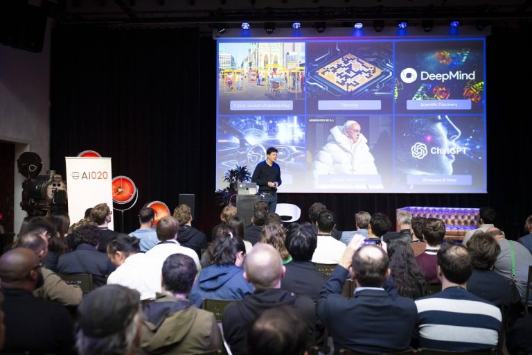 A packed hall, with people seen from behind, watching a presentation on a stage in front of a large screen displaying various images.