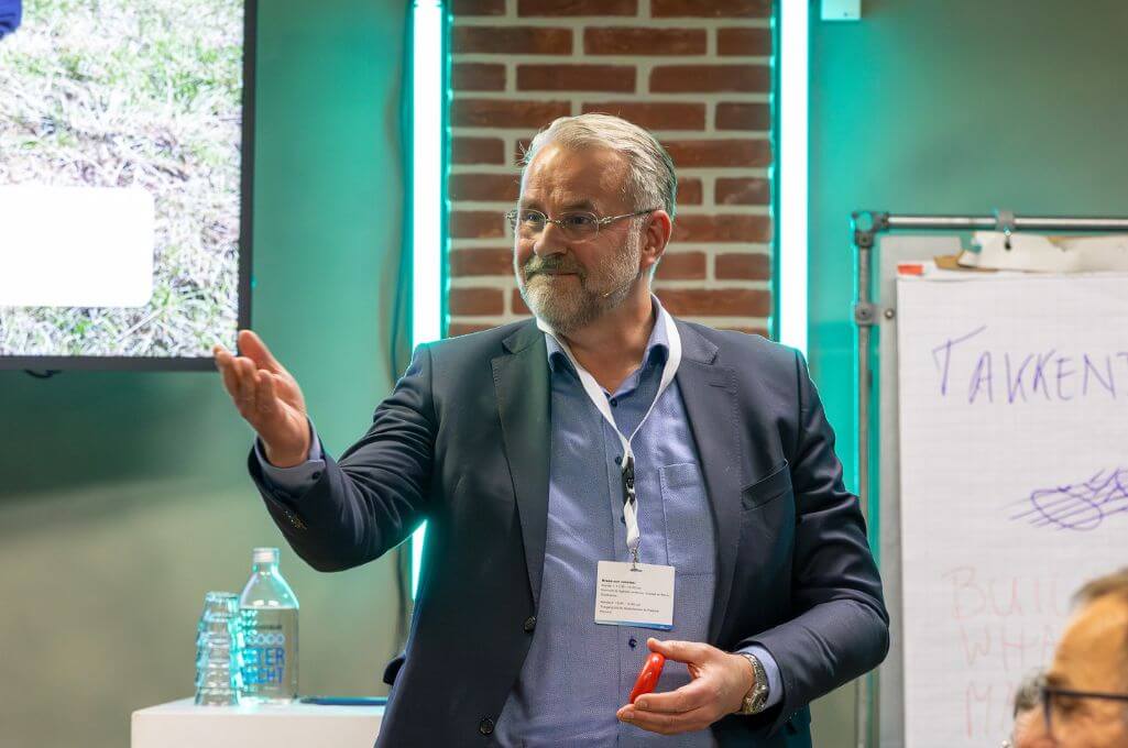 Werkgebied leider Landbouw René Langevoort staat voor een presentatiescherm en nodigt iemand uit om het woord te nemen tijdens de Breakout sessie van Het Nederlandse AI Congres.