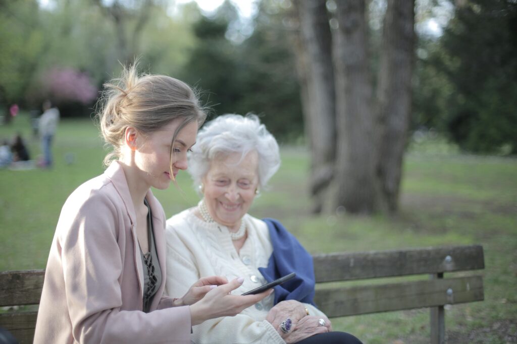 A young woman and an older woman are sitting together on a park bench. The younger woman is holding a smartphone and showing something, while the older woman looks on with a smile. In the background, there are trees and a green, park-like setting.