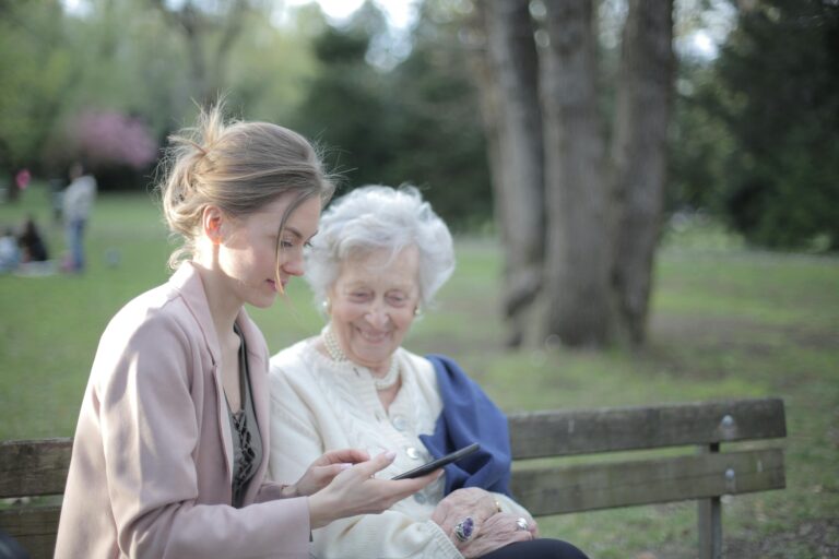 A young woman and an older woman are sitting together on a park bench. The younger woman is holding a smartphone and showing something, while the older woman looks on with a smile. In the background, there are trees and a green, park-like setting.