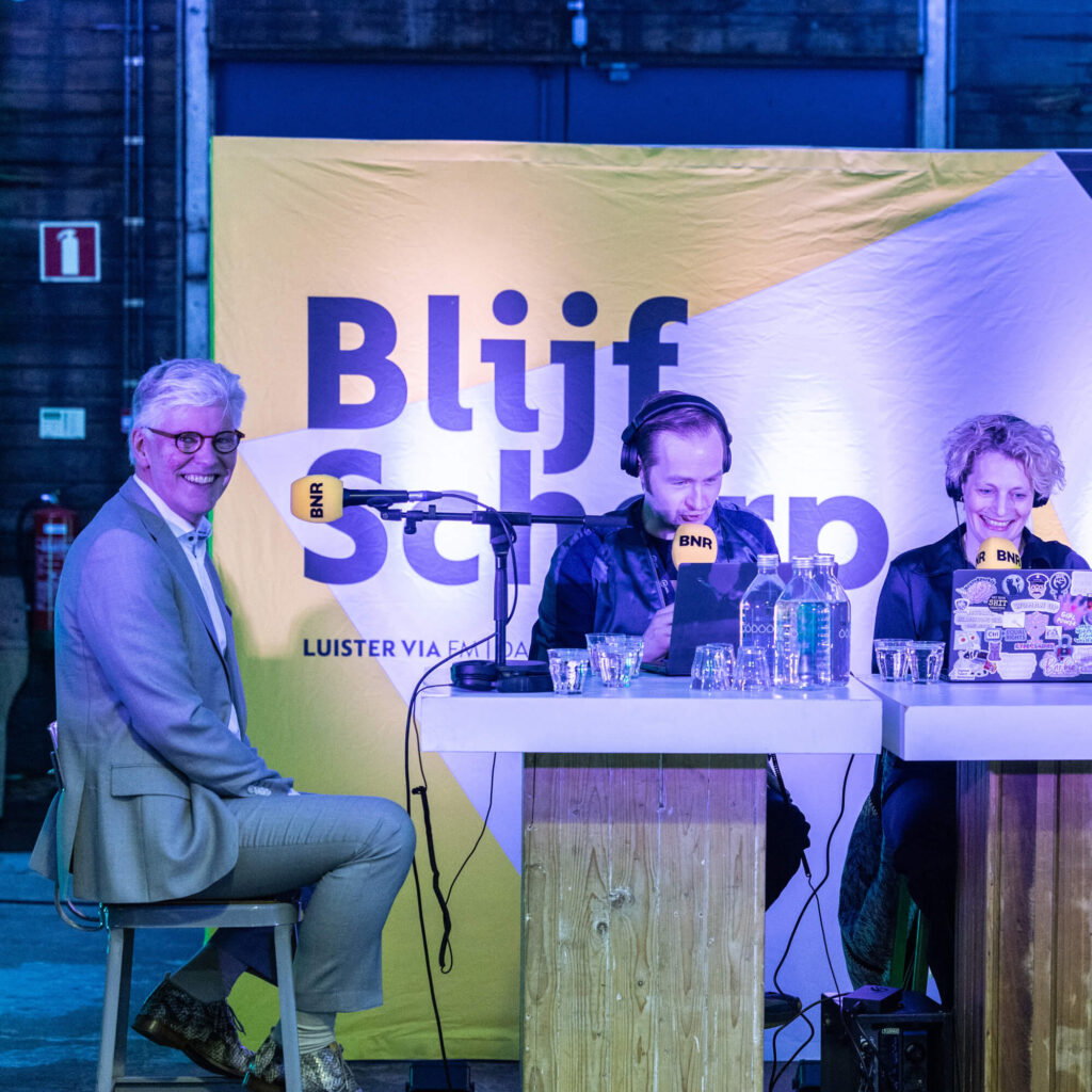 Joe Burik en Lucinda Sterk van De Grote Tech Show en AIC4NL voorzitter Willem Jonker aan tafel met microfoons en geluidsinstallatie tijdens Het Nederlands AI Congres.